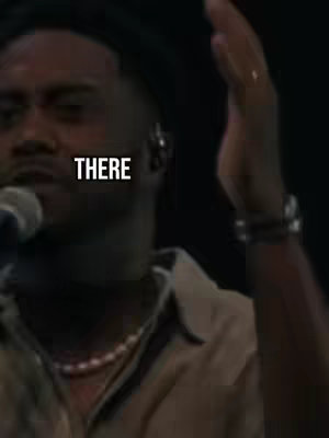 The cover image features a close-up of a Black man with a microphone, possibly singing. The text overlay reads "THERE" in a bold, white font. He wears a neutral-toned shirt and a beaded necklace. The background is dark, suggesting a dimly lit stage or indoor setting. The man’s right hand is raised with his fingers spread, and his arm is visible in the frame. A microphone is in front of his mouth. His face and a portion of his shoulder are in focus.  Accessories like a bracelet and earring are visible.