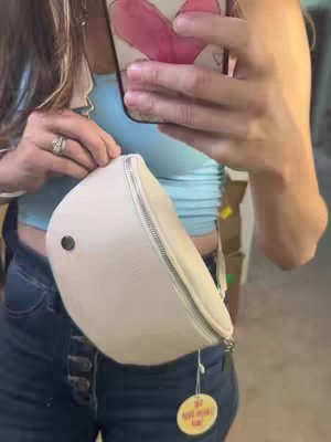 A selfie-style shot of a woman holding a light-colored, oval-shaped bag. The woman is wearing a light blue top and dark blue jeans. Her left hand is holding the bag, and her right hand is holding a phone with a heart-shaped design on the case. A tag is hanging from the bag. The background is blurry, but suggests an indoor setting.