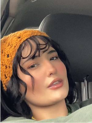 A close-up selfie of a woman in a car, angled slightly upwards. She wears a yellow, crocheted head covering, dark hair with bangs, and has rosy cheeks and glossy lips. The background is out of focus but appears to be the interior of a car with a black seat.