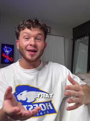 A young man with curly hair smiles and speaks to the camera. He wears a white t-shirt with a blue and yellow logo. The background suggests a domestic setting.