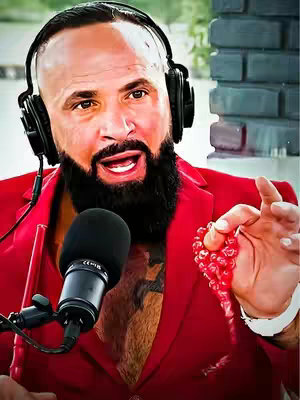 The cover image features a man in a red blazer with a focused expression. He has a bald head, a full black beard, and is wearing headphones around his neck. A microphone is positioned in front of him. In his right hand, he holds a string of red beads. The background appears to be a brick structure.