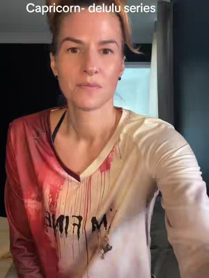 A woman with fair skin and auburn hair is centered in the frame, looking directly at the camera. Her hair is pulled back in a half-up style. She wears a mostly white shirt with a red overlay on one side, and some areas appear to be covered in blood stains. A text overlay at the top reads 'Capricorn- delulu series.'