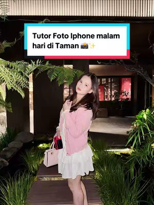 A woman stands outdoors at night in a garden setting, wearing a pink cardigan and white dress, with a small purse. Text overlayed on the image reads 'Tutor Foto Iphone malam hari di Taman'.