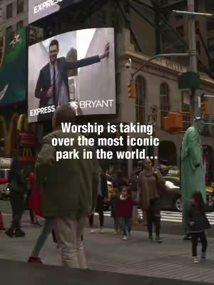 A bustling street scene in New York City, with a large digital billboard displaying a man in a suit. The foreground shows pedestrians walking, including a family with children. The overlay text reads: 'Worship is taking over the most iconic park in the world...'