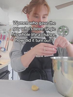 A woman with red hair is shown in a kitchen setting, wearing a gray sweatshirt and a black apron. The text overlay is displayed on the upper part of the picture. She is handling a cookie dough while looking at it.