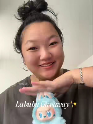 A woman with dark hair pulled back in a bun is smiling and holding a Labubu plush toy in front of her. The words "Labubu Giveaway" are overlaid on the image with twinkling star emojis.