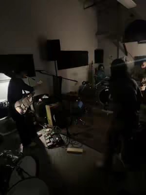 A band is set up and performing in a dimly lit studio. The foreground shows one person playing a guitar, while behind them are drums and other band members. The lighting seems to be focused on the musicians. The tones are dark and moody.