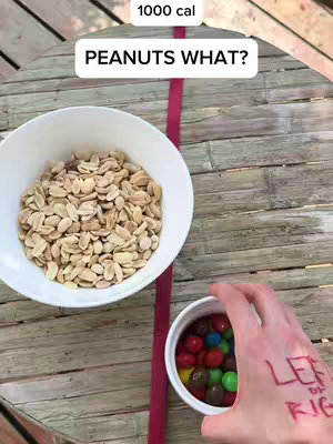 The image shows a top-down view of a round wooden table. On the table are two white bowls; one is filled with peanuts, and the other contains colorful M&M candies. A pink ribbon is stretched across the table between the bowls. A hand is visible, holding the bowl of candies, and has "LEFT or RIGHT" written on the back of it. Above the bowls is a white box with "1000 cal" written in it. Below this text is a white box with "PEANUTS WHAT?" written in it.