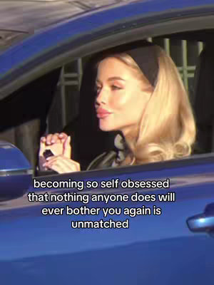 A woman with blonde hair and a black headband is seated in a blue car. She looks slightly to her left with a subtle smirk, while the text overlayed at the bottom shares a message about self-obsession.