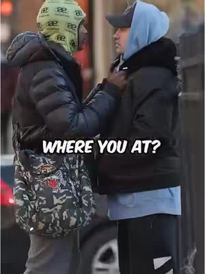 The image shows two men in an urban setting. The man on the left faces toward the right, wearing a black puffer jacket, a patterned bag, and a green and black head covering. The man on the right stands more forward, wearing a black jacket over a blue hoodie and a gray cap. Both men are interacting and facing towards each other. The background suggests a city street scene.