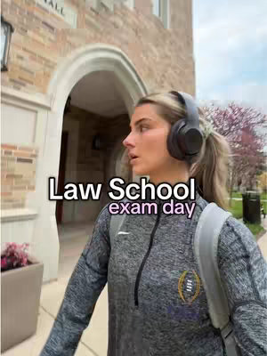 A young woman wearing a gray zip-up jacket, headphones, and a backpack walks towards a grand building entrance. Text overlays read 'Law School exam day'. The background shows a building with arches, and some trees.