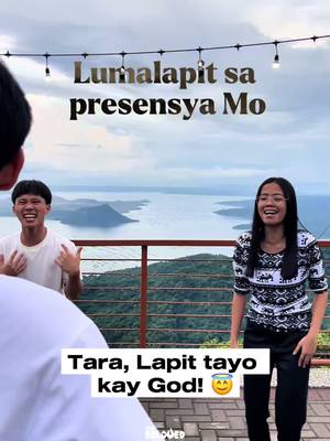 A group of three people is standing outdoors with a scenic background of a lake and mountains under a cloudy sky. The image is taken from a slightly low angle, making the subjects appear larger in the frame. The foreground includes the upper body of a person out of focus. Two individuals are in focus, both smiling. One is a man facing the camera woth his arms raised, wearing a white shirt, and another is a woman with black hair wearing a black and white patterned top. Text overlays are present, including a religious phrase in the background with a call to action. The overall impression is of a joyful, uplifting moment.
