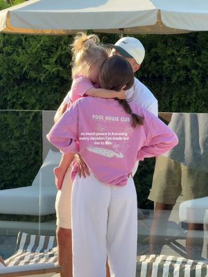 A family of three embracing. A woman in a pink sweatshirt with text on the back that reads: "POOL HOUSE CLUB so much peace in knowing every decision I've made led me to this." She is hugging a small child clad in a pink top standing on the left. A man is embracing the other two from behind. The outdoor setting has green foliage and a patio umbrella in the background.