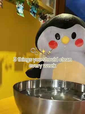 A cover image featuring a large penguin stuffed animal in the center, set against a yellow and white background. The penguin has rosy cheeks and a yellow beak, positioned above a silver bowl filled with water. Text overlay reads '3 things you should clean every week'. Bubbles and stars are added for visual effects.