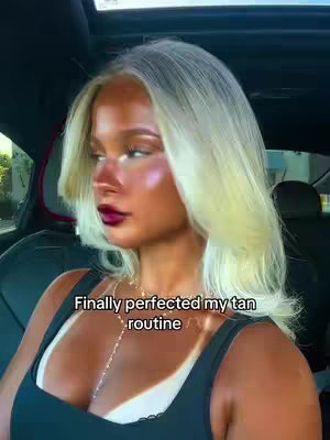 A woman with platinum blonde hair and dark red lipstick sits in a car. The image highlights makeup, tan lines, and a gold necklace. Text overlays read 'Finally perfected my tan routine'.