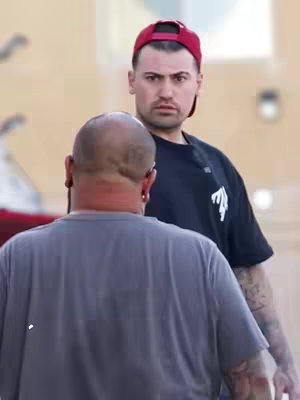 Two men are shown outdoors, likely in an urban setting. One man faces the viewer, wearing a red cap and black shirt. The second man is seen from behind, with a shaved head and a gray t-shirt. The background appears blurred, suggesting a shallow depth of field.