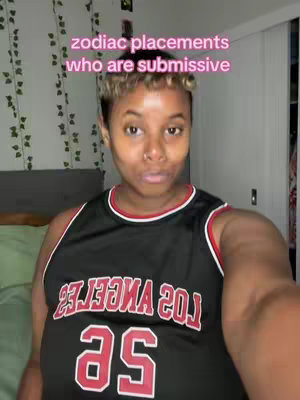 A selfie-style shot of a person wearing a black and red basketball jersey. The person, who appears to be a young adult or adult, is centered in the frame. Above the person's head, text in a pink color is overlaid on the image. The background is blurred, suggesting an indoor setting, with some foliage on the left side.