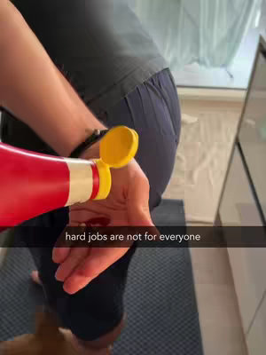 A person is shown from the waist down, wearing dark shorts and a dark t-shirt. They are holding a red ketchup bottle, squeezing what appears to be a small amount of ketchup onto their open palm. The image is taken from a slightly high angle, looking down at the person's hand and lower body. In the background are glimpses of a light-colored wall and a potential window with a light curtain. The text overlay reads 'hard jobs are not for everyone'.