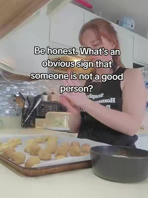 A woman is in a kitchen, with light-colored cabinets in the background. She is wearing a black tank top. In the foreground is a baking tray with several small, dough-like shapes, a black bowl, and an area of the countertop. Text overlays the image, asking a question.