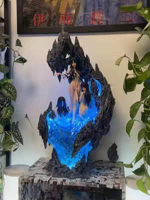 The image features a detailed statue of several figures from the Junji Ito's 'Tomie' series. The statue is centered, taking up most of the frame and has a dark, elaborate structure with a glowing, blue internal light. The statue appears to be sitting on a base with images adhered to it. The background includes a framed piece art and some greenery.