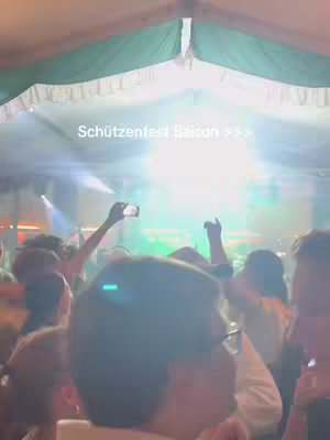 A blurry low-angle shot inside a tent at a crowded event. People are clustered, some holding up phones, with a bright light source at the end of the tent. The focus is on the backs of people's heads.