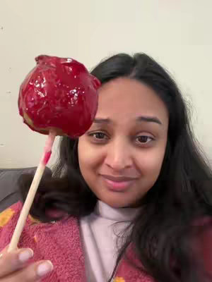 A selfie-style shot featuring a woman holding a large, red candy apple. She's smiling and looking toward the camera. The candy apple dominates the foreground, with the woman's face and upper body filling the rest of the frame. The background is a soft, neutral color, likely an interior wall.