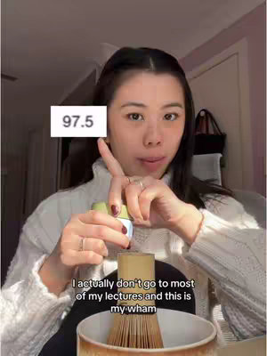 A woman is holding a small green container and pointing her finger to the left of a white box with "97.5" on it. The woman is wearing a white sweater and is positioned to the right of the frame, and a beige bowl with a bamboo whisk inside is in front of her. The background is blurred, with a door and some shadows visible.