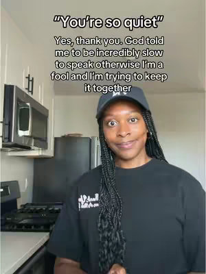 A woman with long braids in a black t-shirt and a black cap is shown from the chest up, in a kitchen setting. Text overlays are centered above her, and she is smiling and looking at the viewer.