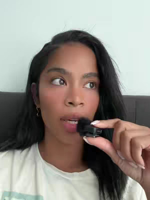 A woman with dark hair holds a small black microphone towards her mouth, looking slightly to the side. She wears a white t-shirt. The background is a light gray wall.