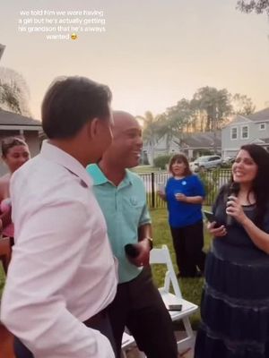 A group of people are gathered outdoors. A man in a light pink shirt faces a man, and another man in a teal shirt is in between them, all looking towards a woman speaking into a microphone, suggesting a presentation or announcement. A woman in a blue shirt stands in the background.