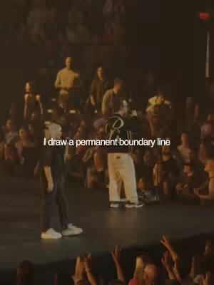A concert stage with two figures in the middle and a crowd in the background. The front figure is wearing a white and black outfit, the second person is wearing a jacket with a 'R' on it and white pants with a black stripe. The stage is dimly lit with overhead lights or spotlights.