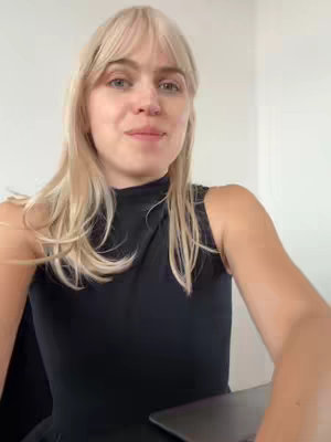 A woman with blond hair and bangs looks at the camera with a soft smile and open expression. She wears a black sleeveless top and her arms are visible. The background is a plain white wall.