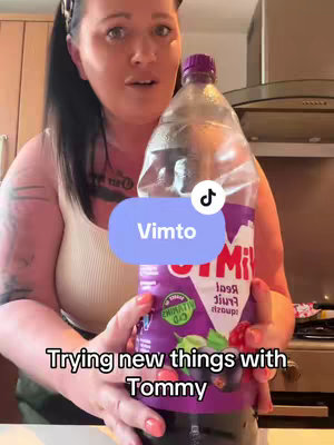 A woman with dark hair pulled back in a ponytail holds a large bottle of Vimto. She is looking at the camera with an open mouth and an interested expression. Text on the bottom of the screen, "Trying new things with Tommy". A branded sticker indicating Vimto. The image is taken in front of a kitchen setting.