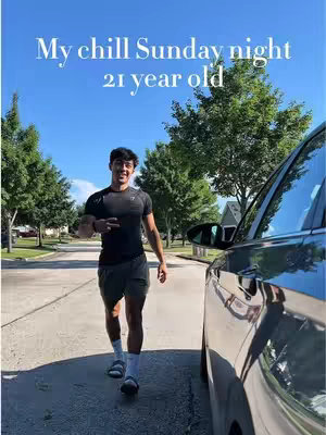 A young man in athletic wear walks towards the camera on the sidewalk, with a car parked on the right side. The sky is blue with some trees in the background.
