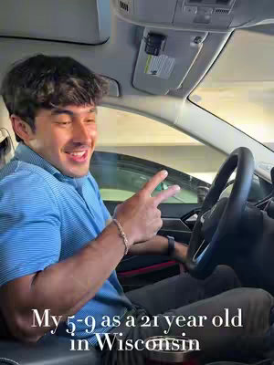 A young man is seated inside a car, likely the driver's seat, with a bright, sunny background visible through the windows. He has short, dark hair and is smiling, making a peace sign with his fingers. The interior of the car is visible, including the steering wheel and part of the dashboard.