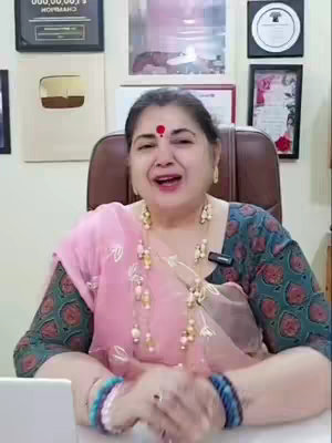 A woman with a red bindi sits in a chair, smiling and talking. She wears a pink scarf over a patterned top and has long necklaces and bracelets. The background has framed certificates and awards.