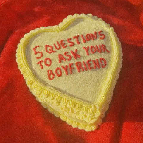 The cover image features a heart-shaped cake decorated with yellow frosting and a decorative border. The text '5 QUESTIONS TO ASK YOUR BOYFRIEND' is piped in red frosting across the center of the cake. The cake is placed on a red fabric background, creating a warm and inviting visual.