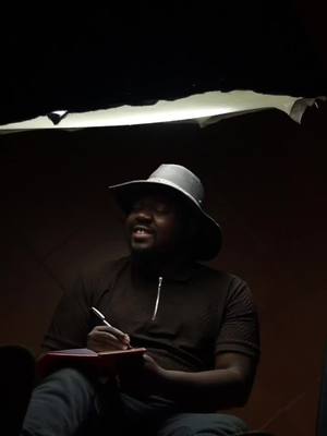 A man wearing a hat is seated and holding a pen and a notebook. The lighting is focused on the man. The background is dark.