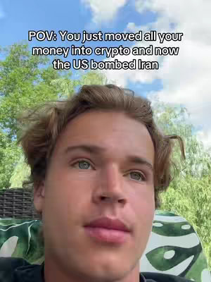 A young man with fair skin and light brown, curly hair is centered in the frame. The background is a blurred mix of green foliage and a cloudy sky. Text in a white box overlaying the image reads: 'POV: You just moved all your money into crypto and now the US bombed Iran'. The person in the photo is wearing a dark-colored shirt, and the lighting is soft, suggesting a daylight setting.
