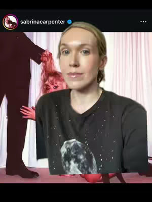 The image is a selfie-style video. The main focus is on the creator, a person with fair skin and light-colored hair, wearing a black shirt. The creator is centered in the frame. In the background, a second person with pink hair is visible, partially obscured behind the creator. The background has a blurry, pinkish cast with what appears to be a draped white fabric behind the figures. The overall composition suggests a candid, possibly humorous video.