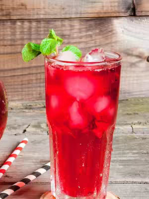 The image features a vibrant red drink in a tall glass with ice cubes and a sprig of mint on top, set against a wooden background. A straw with red and white stripes and a black and white straw are visible on a wooden surface near the glass, adding visual interest with their patterns and colors. The composition is well-lit, highlighting the freshness of the drink.