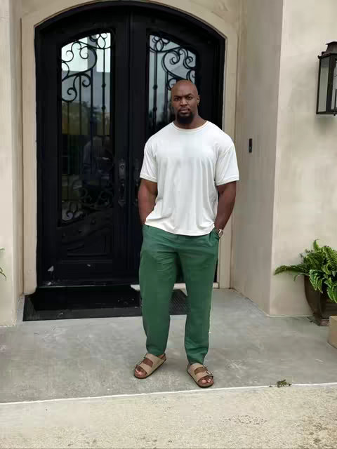 A full-body shot of a man standing in front of a double-door entrance. He is wearing a white t-shirt, green pants, and sandals. He stands with his hands in his pockets, in front of a black door with ornamental ironwork. The backdrop is a house exterior, and to the side, there is a lantern and a potted plant.
