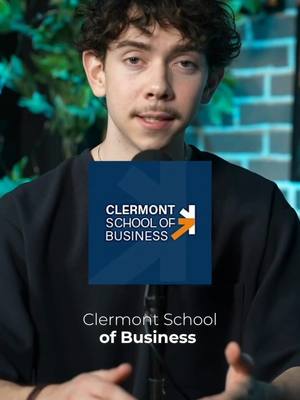A young man with curly hair and a mustache is centered in a video, against a background of green foliage and a brick wall. He wears a black shirt and speaks into a microphone behind a branded logo that says 'Clermont School of Business'.