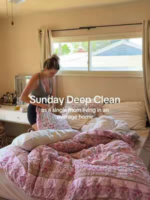 A woman in a casual outfit is in a bedroom arranging pillows. The room is well-lit with natural light coming through a window. The bed has a floral patterned coverlet, and pillows are scattered around. The overlay text says, "Sunday Deep Clean as a single mom living in an average home."