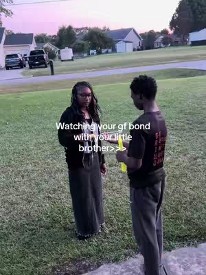 A video cover shows a grassy area in front of houses. In the foreground, a man and a woman stand facing each other. The man on the right, holding a yellow bubble wand, has a black t-shirt and gray pants. The woman wears a black jacket and skirt. Text overlayed on the image reads 'Watching your gf bond with your little brother >>'.