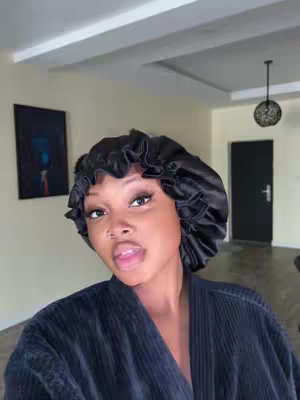 A selfie-style shot featuring a person against an interior backdrop. The person is wearing a black satin bonnet and a dark robe. The setting includes a door on the right, a painting on the left, and a pendant light. The overall color scheme is dark, with varying shades of black and neutral tones.