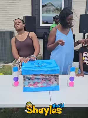 The cover image shows two women standing behind a table with a blue box filled with colorful items. The box is centered on the table, flanked by decorated bottles. The women are smiling and appear to be engaged in a game or activity. A child is standing to the right of the women. The background includes a house and some outdoor equipment, suggesting a backyard setting.