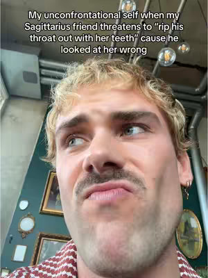 A man with blonde hair and a mustache looks up with a mix of confusion and suspicion on a background with a wall of framed paintings.