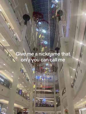 An upward shot of a large, multistoried interior space, likely a shopping mall or a similar public building. The architecture features multiple levels with balconies visible on either side, leading up towards a high ceiling. Hanging from the ceiling are elaborate decorations, possibly for the holiday season, including strings of lights, snowflake ornaments, and other festive elements. The image is well-lit, with the light reflecting off the clean, white surfaces of the building, creating a bright and open atmosphere. The overall composition emphasizes vertical lines and symmetry, drawing the eye towards the decorative elements that add color and vibrancy to the scene.
