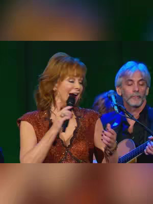The cover image shows a musical performance on stage. Reba McEntire, a woman with red hair, sings into a microphone, wearing a brown sequined top with a V-neck. Another person is to the right with a guitar. Others are visible in the background, suggesting a live setting. Lighting is warm, and the overall composition focuses on the performance.
